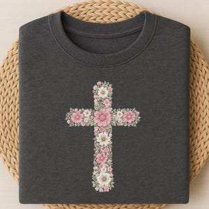 May include: A dark charcoal gray crew neck sweatshirt featuring a central graphic of a cross composed of various pink and white flowers with green leaves. The floral arrangement includes detailed petals and foliage in soft pastel tones.