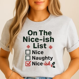 May include: A light grey t-shirt featuring the text "On The Nice-ish List" in dark green. The design includes a checklist with "Nice", "Naughty", and "Nice-ish" options, with "Nice-ish" checked in red. Festive asterisks and holly accents complete the design.