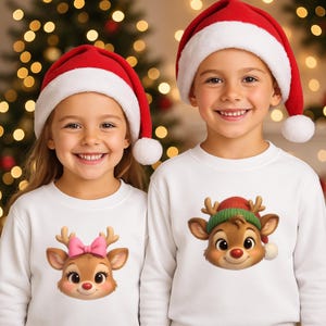 May include: Two children wearing white sweatshirts with reindeer graphics and red Santa hats with white trim and a white pom-pom. The reindeer graphics have a pink bow and a green hat. The background is a blurred Christmas tree.