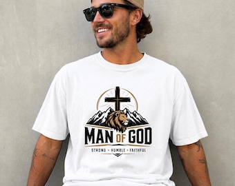 May include: A white short-sleeved t-shirt featuring a graphic print of a lion head, mountain range, and a cross. The text reads MAN OF GOD with the words STRONG, HUMBLE, FAITHFUL underneath. The design uses black, brown, and gold color tones.