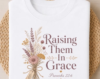 May include: A white t-shirt featuring a graphic of a dried wildflower bouquet tied with a string and a small tag. The text reads "Raising Them In Grace" in a dark purple serif font, followed by a small heart and the citation "Proverbs 22:6".