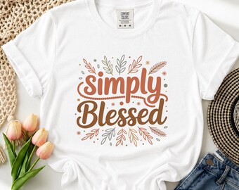 May include: A white short-sleeve t-shirt featuring the text "simply Blessed" in brown and orange script lettering, surrounded by a decorative botanical graphic of leaves and small stars in muted earth tones, with a Comfort Colors brand label.