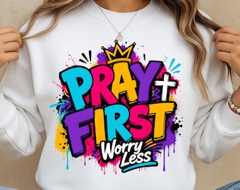 May include: A white crewneck sweatshirt featuring a colorful graphic print with the text PRAY FIRST Worry Less. The design includes a yellow crown, a white cross, and vibrant paint splatter accents in shades of blue, pink, purple, and yellow.