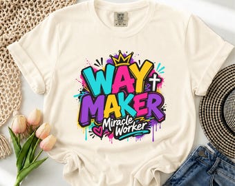 May include: A cream-colored short-sleeve t-shirt featuring a vibrant graphic print with the text "WAY MAKER" in large, colorful block letters, "Miracle Worker" in script, a yellow crown, a white cross, and a small pink heart, all accented by paint splatters.