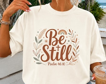 May include: A cream-colored short-sleeve t-shirt featuring a graphic print with the text Be Still and Psalm 46:10. The design includes a brown cross, a small rainbow, sun icons, and stylized leafy branches in muted tones of brown, green, and orange.