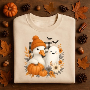 May include: Cream-colored sweatshirt featuring a whimsical Halloween design. The graphic includes a duck wearing an orange hat, a friendly ghost, pumpkins, and flowers. Two black bats fly above the characters. The sweatshirt is surrounded by fall-themed decorations.