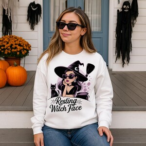 May include: White sweatshirt featuring a witch graphic with sunglasses, a hat, and a black cat. The text "Resting Witch Face" is below the graphic. Ideal for Halloween.