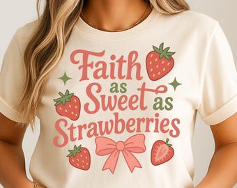 May include: Cream-colored t-shirt with the text "Faith as Sweet as Strawberries" in pink and green. The design includes strawberry illustrations and a pink bow. The shirt has short sleeves and a crew neck.