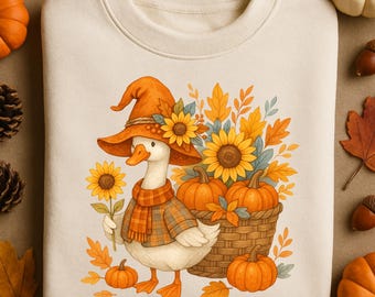 Op de afbeelding: Crèmekleurige sweatshirt met een grillige illustratie van een gans met een heksenhoed, sjaal en geruite jas. De gans houdt een zonnebloem vast en is omringd door pompoenen, zonnebloemen en herfstbladeren. Er is ook een mand met pompoenen.