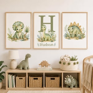 Puede incluir: Tres cuadros de pared con ilustraciones en acuarela de dinosaurios verdes en un entorno de selva. El cuadro central muestra una letra H grande y el nombre Hudson. Los cuadros están colocados sobre una unidad de almacenamiento de madera.