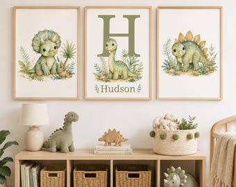 May include: Three framed wall art prints featuring watercolor illustrations of green dinosaurs in a jungle setting. The center print displays a large letter H and the name Hudson. The prints are displayed above a wooden storage unit with baskets.