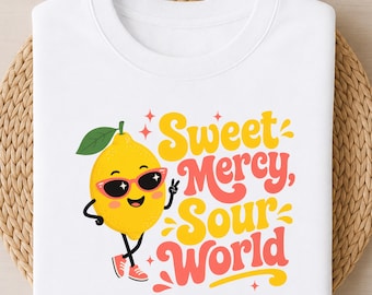 May include: A white t-shirt featuring a graphic of a yellow lemon character wearing pink sunglasses and red sneakers. The lemon is making a peace sign with one hand. The text "Sweet Mercy, Sour World" is printed in yellow and pink wavy lettering.