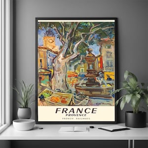 Op de afbeelding: Een ingelijste vintage reisaffiche voor Frankrijk, Provence, door de Franse Spoorwegen. Het kunstwerk toont een fontein, gebouwen en een grote boom op een stadsplein. De poster heeft een crèmekleurige rand met de woorden "FRANCE" en "PROVENCE" in vette tekst.