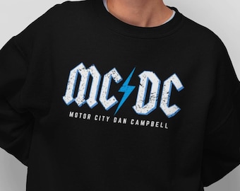 May include: Black sweatshirt with a white and blue graphic design that says "MC/DC Motor City Dan Campbell".
