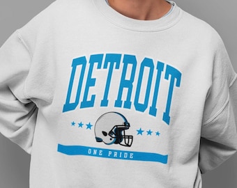 May include: White sweatshirt with the word "DETROIT" in large, light blue block letters. Below the word "DETROIT" is a football helmet graphic and the words "ONE PRIDE" in light blue. The sweatshirt is a crew neck.