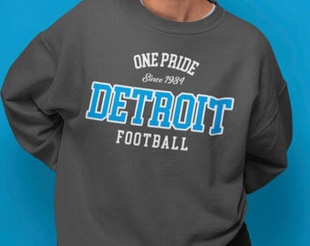 May include: Dark gray sweatshirt with the text "ONE PRIDE Since 1934 DETROIT FOOTBALL" in white and blue. The sweatshirt is a crew neck style and is made of a soft material. The background is a solid blue color.