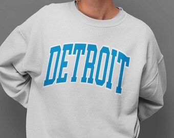 May include: A white sweatshirt with the word "DETROIT" in large, arched, light blue letters. The text has a darker blue outline. The sweatshirt is a crew neck style and appears to be made of a soft material.