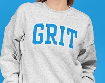 May include: A light gray sweatshirt with the word "GRIT" in bold, blue, block letters across the chest. The sweatshirt has a crew neck and long sleeves. The background is a solid blue.