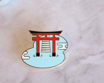May include: A gold-colored enamel pin featuring a red torii gate over a blue body of water with white clouds. The torii gate has black accents and a white staircase.