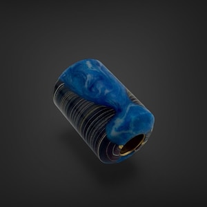 May include: A cylindrical bead featuring a dark, finely lined black and grey base that transitions into a smooth, marbled section of vibrant blue and white resin. The bead has a hollow centre with a metallic gold-toned inner lining.