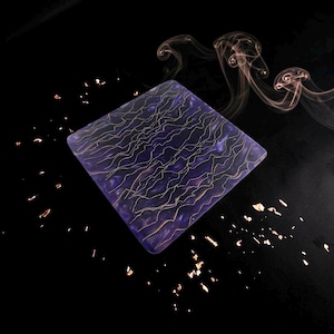 Pode incluir: Um porta-copos quadrado com uma base de mármore roxo profundo e linhas finas e onduladas de cor dourada na superfície. O porta-copos está rodeado por flocos dourados dispersos e volutas de fumo contra um fundo escuro.