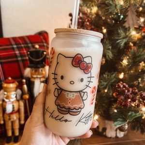 May include: A frosted glass tumbler with a bamboo lid and a Hello Kitty design. The tumbler is filled with a light brown beverage and has a straw sticking out of the top. The design features Hello Kitty dressed as a gingerbread person with candy cane accents. The text "Hello Kitty" is written on the tumbler.