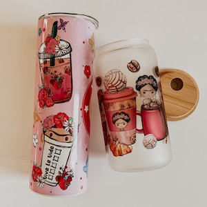 May include: Two colorful, reusable tumblers with lids. One tumbler features a pink and red floral design with the text "Viva la Vida". The other tumbler features a white background with a coffee cup, a pastry, and a cartoon woman with a flower crown.