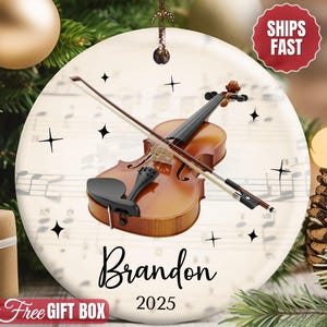 May include: A round ceramic ornament featuring a brown violin and bow centered over a background of musical sheet notes. The ornament includes the name Brandon and the year 2025 printed in black text, accented by small black star icons.