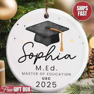 May include: White ceramic graduation ornament featuring a black mortarboard with a gold tassel. The ornament is personalized with the name "Sophia", "M.Ed. Master of Education USC 2025". Decorative stars and a graduation theme.