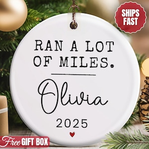 May include: White ceramic Christmas ornament with the text "RAN A LOT OF MILES. Olivia 2025" in black. A small red heart is at the bottom. The ornament is hanging on a Christmas tree with a gold ornament and a pine cone.