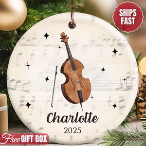 May include: A round, cream-colored Christmas ornament featuring a watercolor illustration of a cello and bow. The ornament has musical notes in the background, with the name "Charlotte" and the year "2025" printed below the cello.
