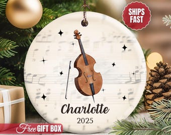 May include: A round white ceramic ornament featuring a brown double bass and bow illustration centered over a musical staff background. The name Charlotte is printed in black script below the instrument, with the year 2025 underneath.