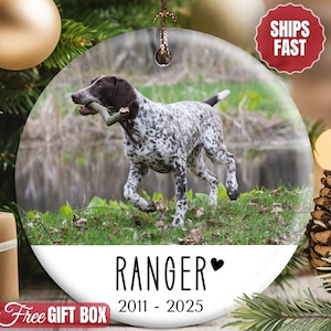 May include: A round, clear glass ornament featuring a German Shorthaired Pointer dog with a stick in its mouth. The ornament has the name "RANGER" and the years "2011 - 2025" printed on it. The background includes greenery and a blurred water scene.