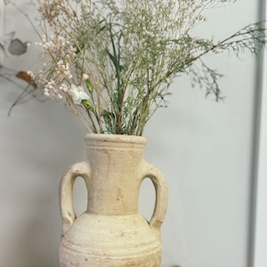 May include: A beige, antique-style vase with two handles, filled with dried flowers and greenery. The vase has a textured surface and a rounded shape, sitting on a wooden surface. The arrangement includes white and beige flowers.