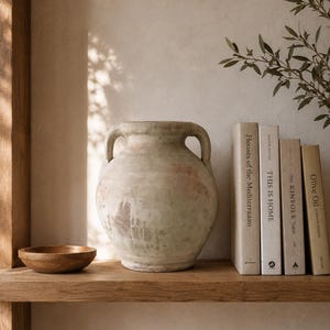 May include: A weathered, light-toned ceramic amphora vase with two side handles, positioned next to a small wooden bowl and a row of four books titled Houses of the Mediterraaan, THIS IS HOME, The Kinfolk Table, and Olive Oil on a wooden shelf.
