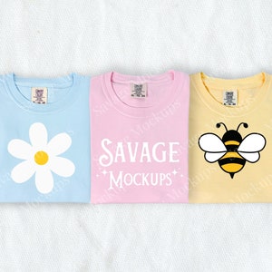 May include: Three folded t-shirts with different designs. The first shirt is light blue with a white daisy. The second shirt is pink with the text "SAVAGE MOCKUPS". The third shirt is yellow with a cartoon bee.