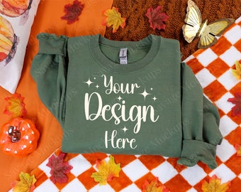 May include: A green sweatshirt with the text "Your Design Here" on the front. The sweatshirt is lying on a checkered orange and white blanket with fall leaves scattered around it.