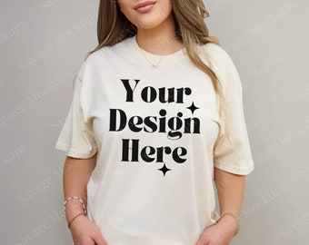 May include: Cream-colored oversized t-shirt with the text "Your Design Here" in bold black letters. The model is wearing blue jeans. The shirt has short sleeves and a relaxed fit, perfect for showcasing custom designs.