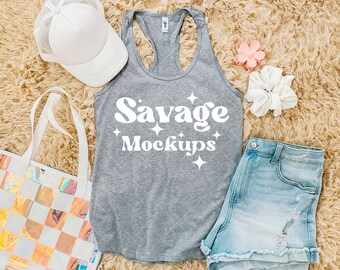 Op de afbeelding: Een grijs tanktop met de tekst "Savage Mockups" in wit. Het tanktop ligt op een lichtbruin, pluche oppervlak met een witte mesh baseballpet, een witte scrunchie, een lichtblauwe denim short en een roze scrunchie.