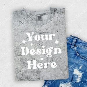 May include: A gray t-shirt with a distressed, textured look. The shirt has the text "Your Design Here" printed in white on the front. The shirt is folded and laid on a white surface with a pair of blue jeans.