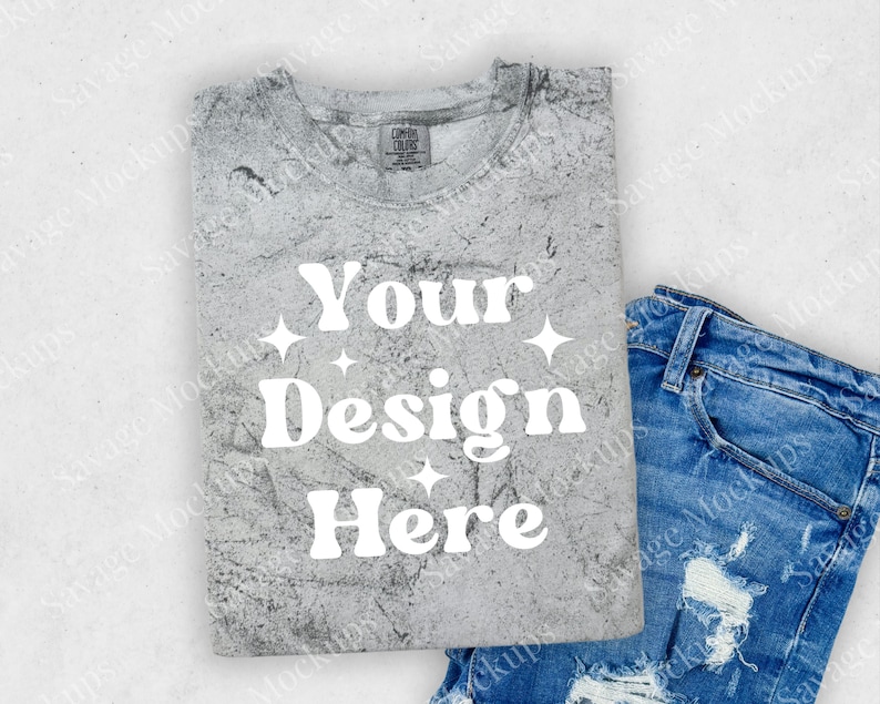 May include: A gray t-shirt with a distressed, textured look. The shirt has the text "Your Design Here" printed in white on the front. The shirt is folded and laid on a white surface with a pair of blue jeans.