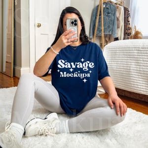 May include: A woman wearing white pants and white sneakers is sitting on a white rug. She is holding a phone in front of her face. She is wearing a navy blue t-shirt with the text "Savage Mockups" in white.