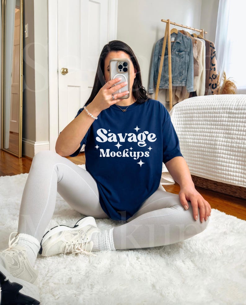 May include: A woman wearing white pants and white sneakers is sitting on a white rug. She is holding a phone in front of her face. She is wearing a navy blue t-shirt with the text "Savage Mockups" in white.