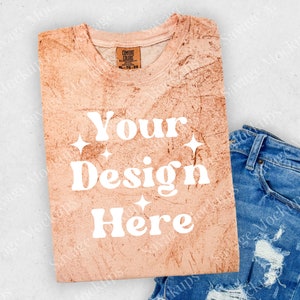 May include: A light peach-colored t-shirt with a distressed look. The shirt has the text "Your Design Here" in white with a retro font and small white stars. The shirt is folded and laid on a white surface with a pair of blue jeans.