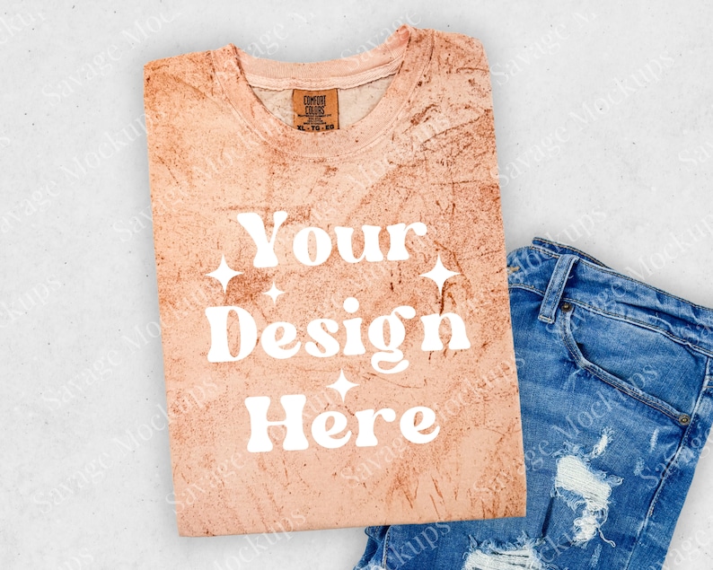 May include: A light peach-colored t-shirt with a distressed look. The shirt has the text "Your Design Here" in white with a retro font and small white stars. The shirt is folded and laid on a white surface with a pair of blue jeans.