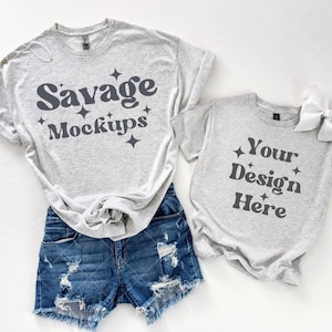 Puede incluir: Dos camisetas grises de manga corta. La camiseta más grande tiene el texto "Savage Mockups" impreso en ella. La camiseta más pequeña tiene el texto "Your Design Here" impreso en ella.