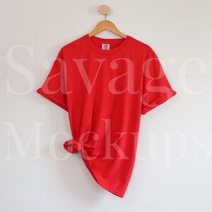 May include: Red t-shirt hanging on a wooden hanger against a white wall. The shirt is knotted at the bottom.