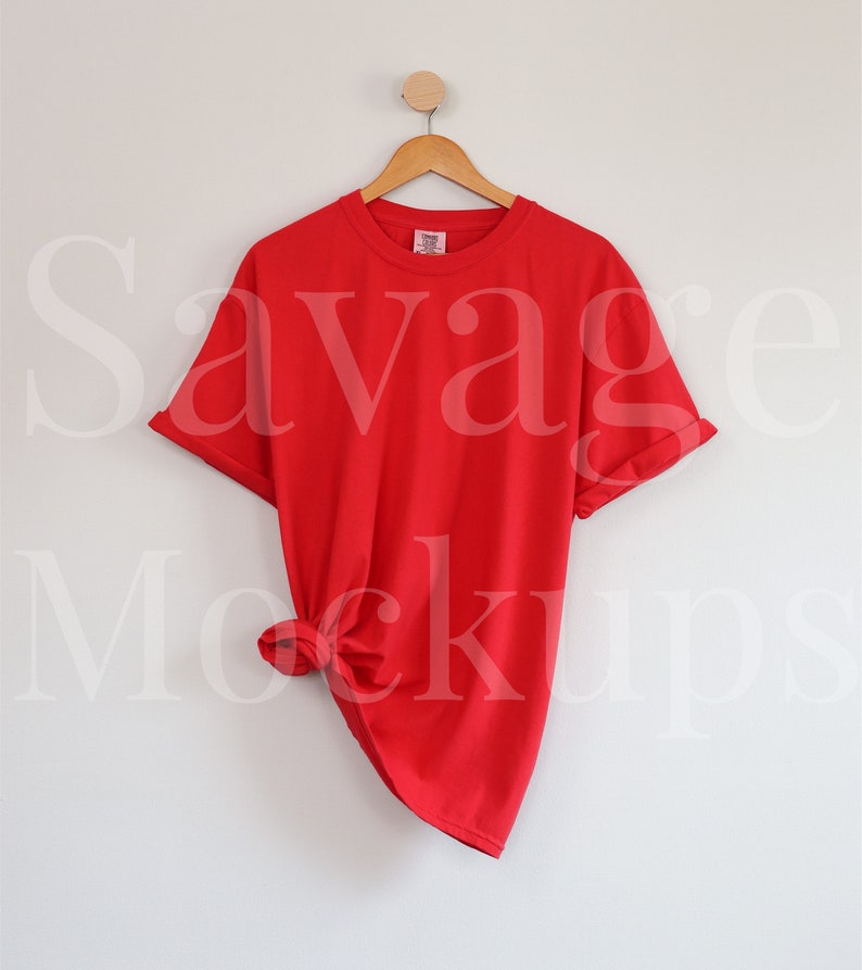 May include: Red t-shirt hanging on a wooden hanger against a white wall. The shirt is knotted at the bottom.