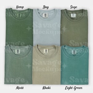 May include: Six folded t-shirts in different shades of green, including hemp, bay, sage, moss, khaki, and light green. Each shirt has a small tag with the words "Comfy Colors" on it.