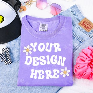 May include: A purple t-shirt with the text "YOUR DESIGN HERE" in white retro font. The shirt is decorated with two white daisy flowers. The shirt is laid flat on a white surface with a pair of blue jeans, a pink scrunchie, and a black and white checkered hair clip.